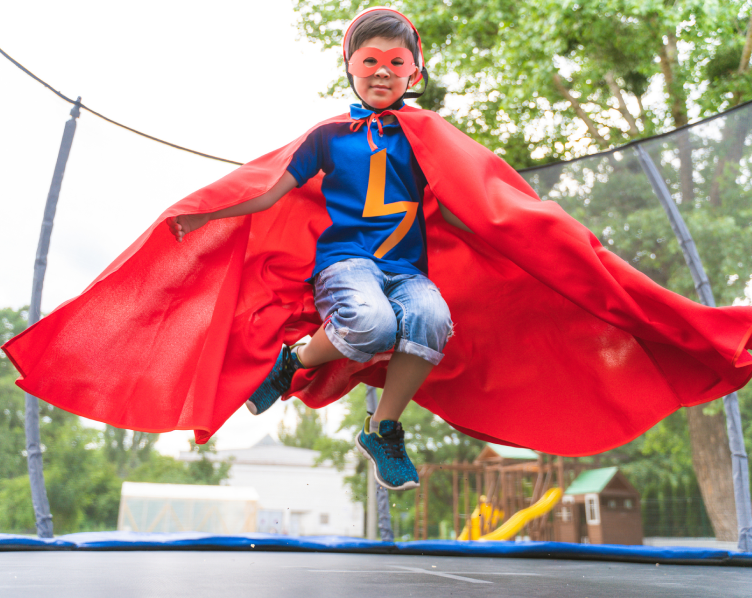 A boy in a superman costume jumping on a trampoline.